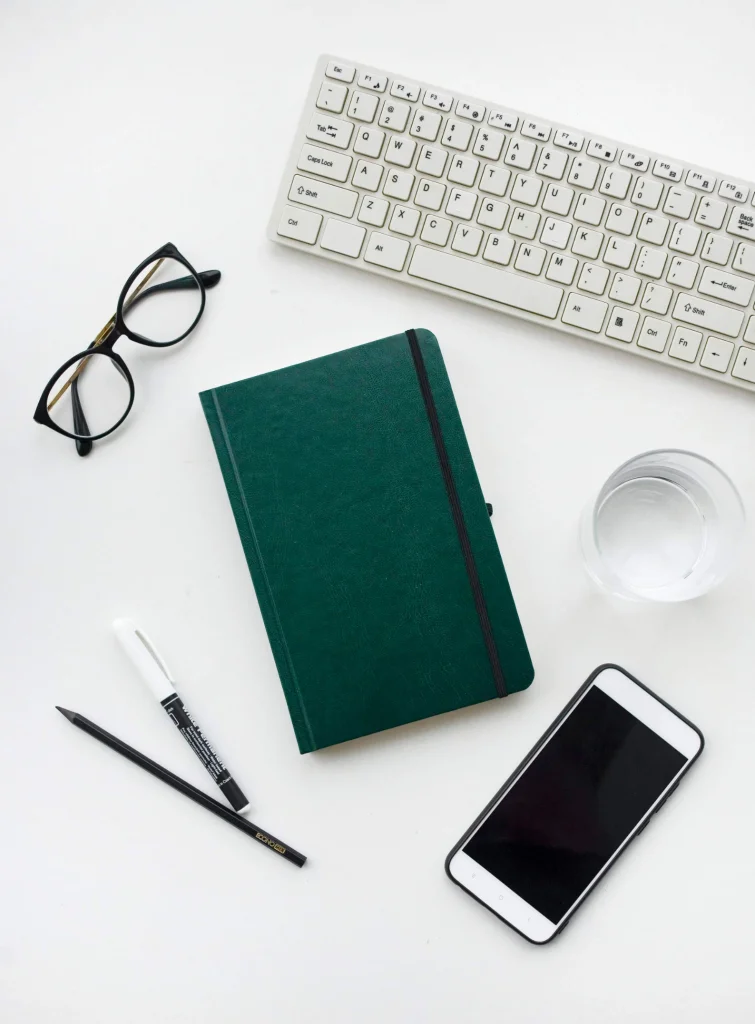 Białe biurko na którym znajduje się klawiatura, okulary, notes, pisak, ołówek, szklanka i telefon komórkowy.