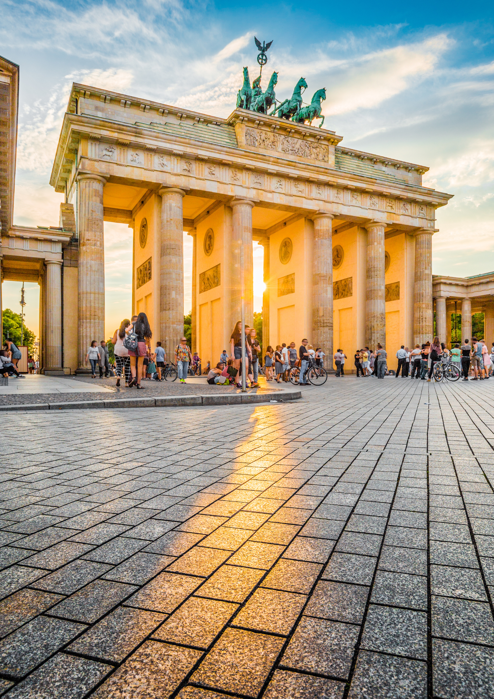 Brama Brandenburska w Berlinie o zachodzie słońca, z kolumnami w stylu klasycystycznym i kwadrygą na szczycie. Na placu przed monumentem spacerują turyści, a promienie słońca odbijają się od kamiennej nawierzchni.