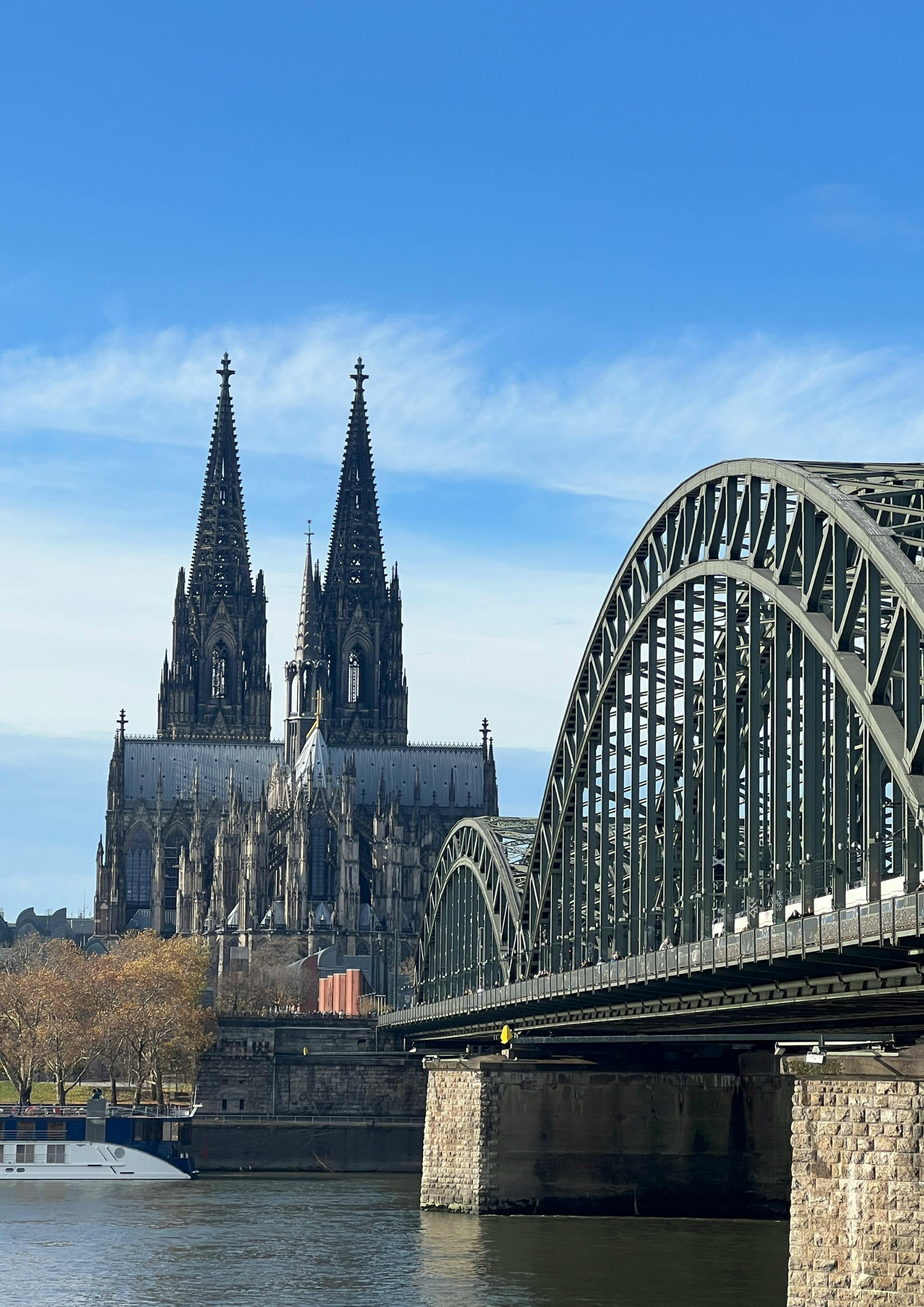 Katedra Kolońska z dwiema strzelistymi wieżami widziana zza mostu Hohenzollernów nad rzeką Ren, w słoneczny dzień z błękitnym niebem.