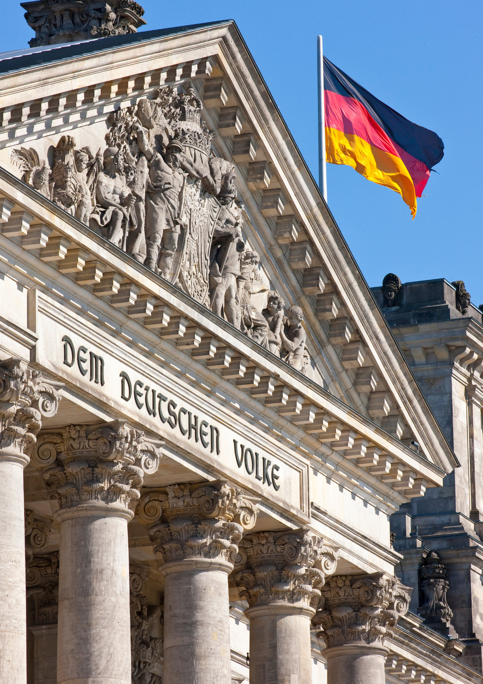 Fragment fasady budynku Reichstagu w Berlinie z kolumnami i rzeźbionym frontonem z napisem „Dem Deutschen Volke”, nad którym powiewa niemiecka flaga na tle błękitnego nieba.