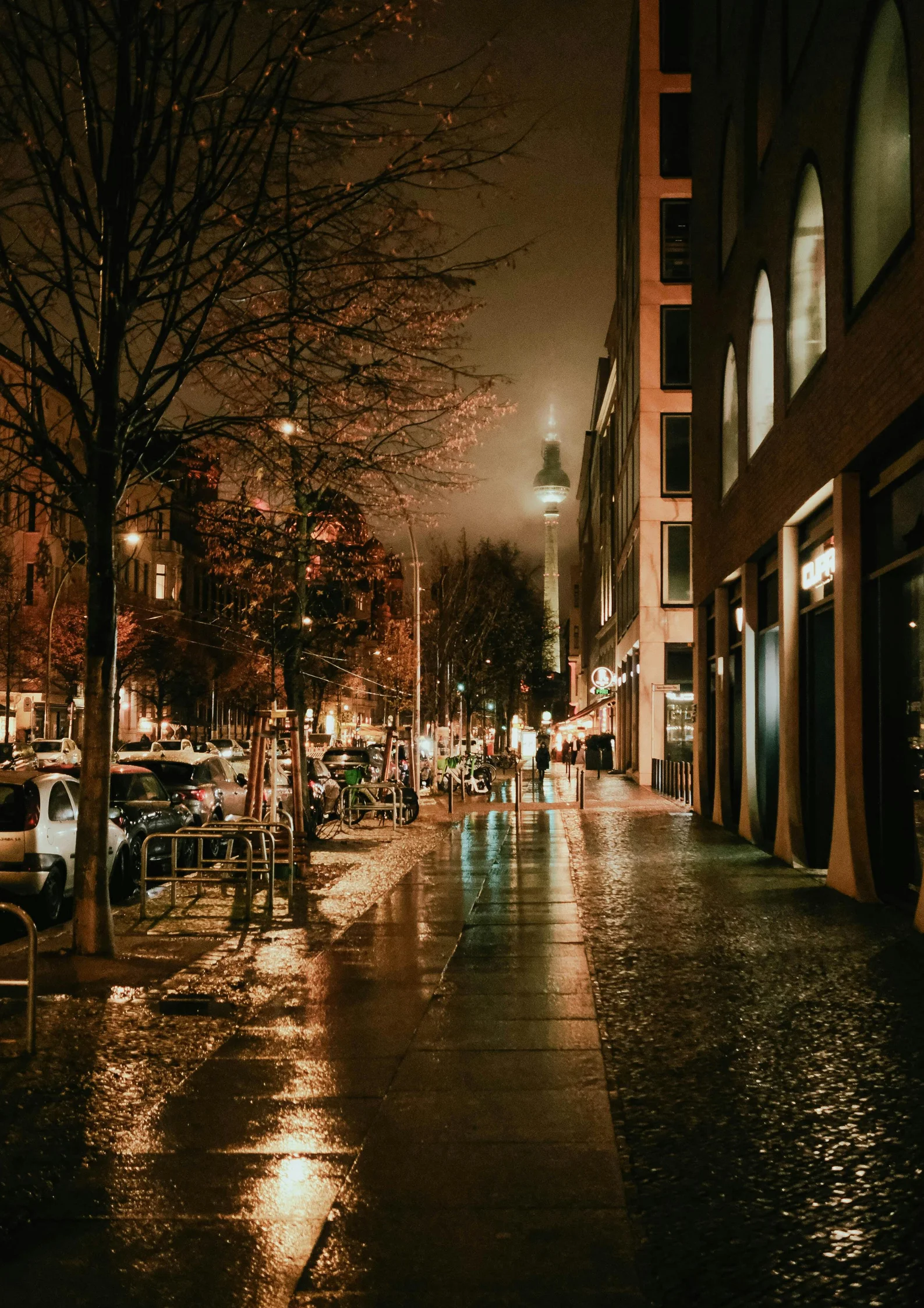 Mokra, oświetlona latarniami ulica w Berlinie nocą, z zaparkowanymi samochodami i drzewami, w oddali widoczna wieża telewizyjna Fernsehturm spowita lekką mgłą.